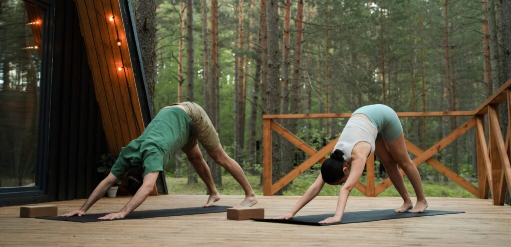 A man and a woman doing yoga on the balcony of a cabin in the woods, surrounded by nature and fresh air.