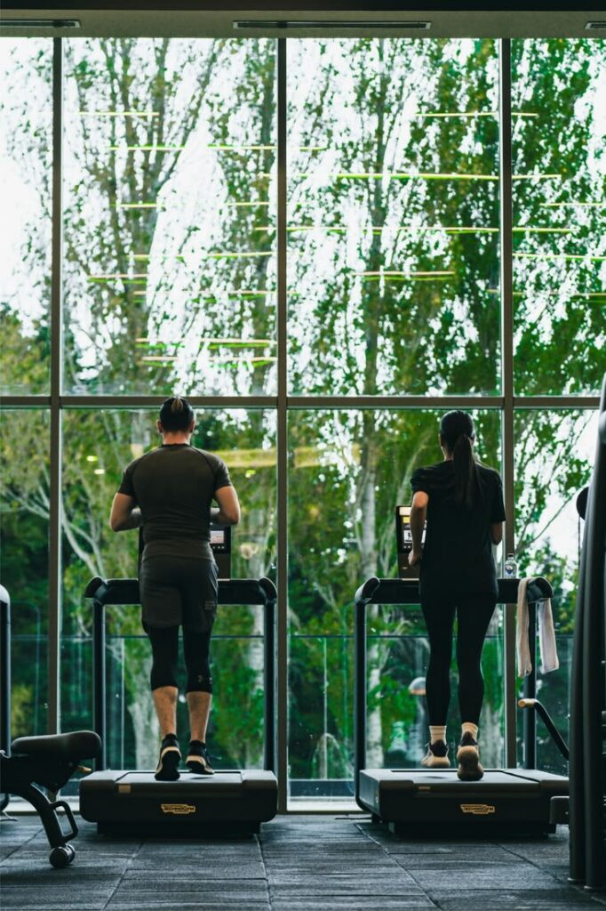 Two adults running on treadmills in front of a large window.