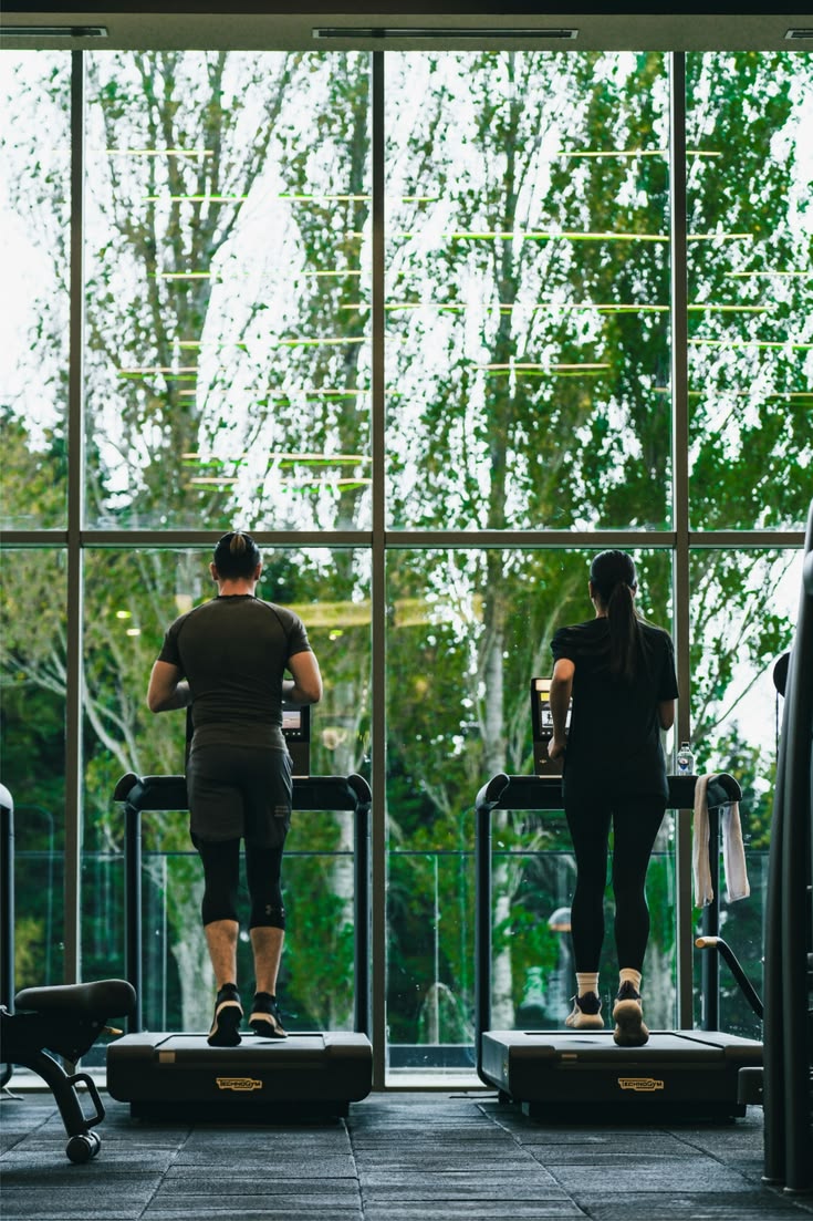 Two adults running on treadmills in front of a large window.
