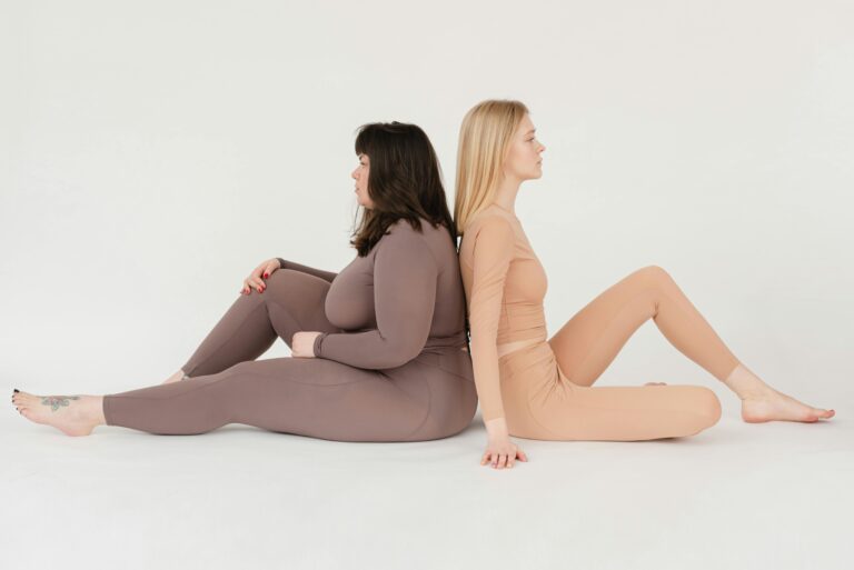 Two women wearing yoga outfits, sitting on the floor back to back, in a relaxed and comfortable pose.