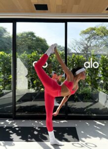 Woman wearing a bright pink Pilates workout set stretching on a mat in a sunlit studio.