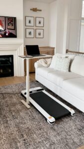 Walking pad positioned under a standing desk with a laptop in a stylish home office and living space.