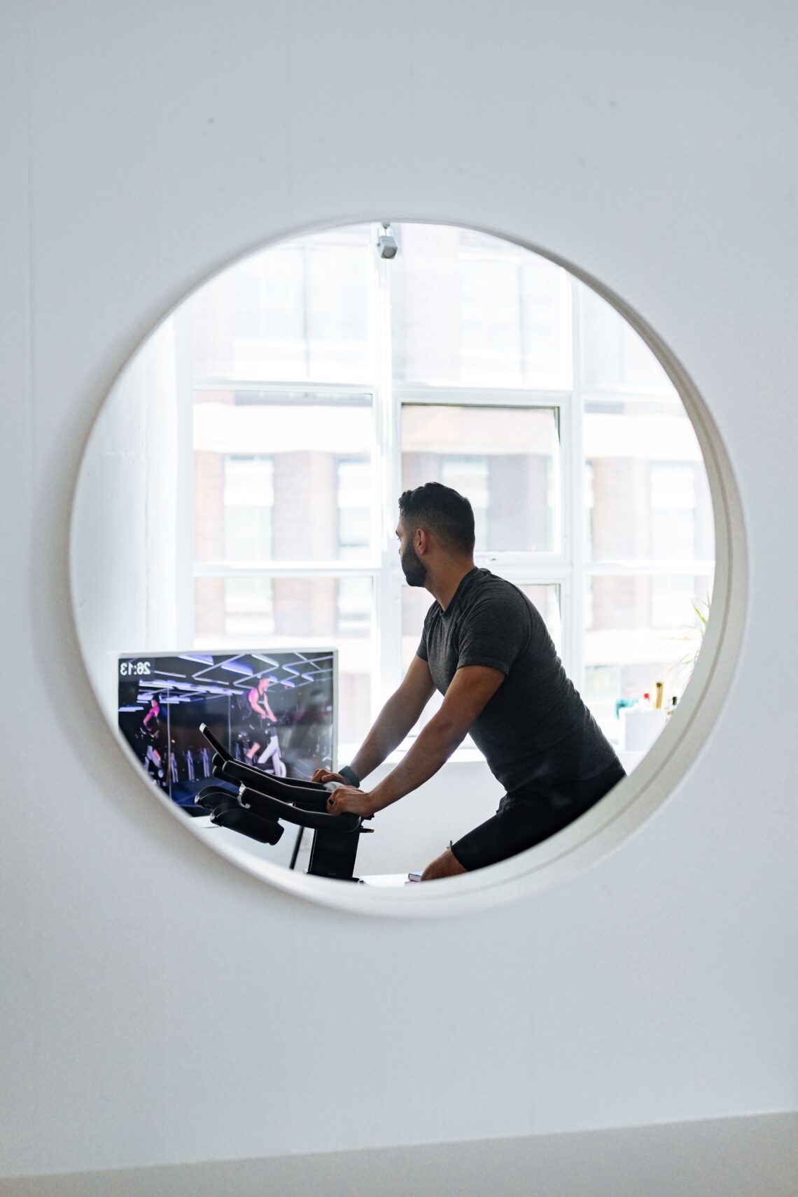 Unrecognizable male in sportswear looking away and reflecting in small round mirror on wall while training on exercise machine in cozy apartment