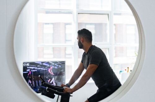 Unrecognizable male in sportswear looking away and reflecting in small round mirror on wall while training on exercise machine in cozy apartment