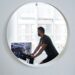 Unrecognizable male in sportswear looking away and reflecting in small round mirror on wall while training on exercise machine in cozy apartment