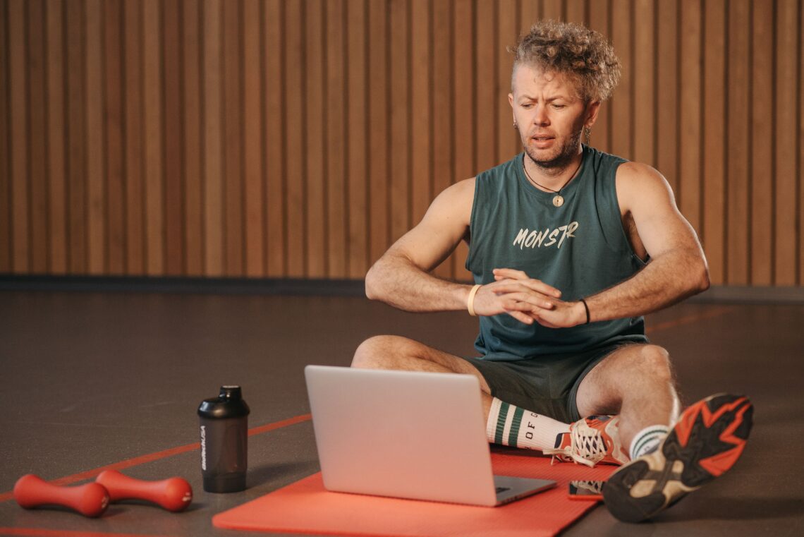 Adult man exercising indoors with a laptop for virtual community cardio fitness training, sitting on a red mat.