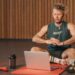Adult man exercising indoors with a laptop for virtual community cardio fitness training, sitting on a red mat.