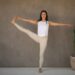 popular 2026 fitness influence Adrienne mishler wearing beige leggings and white t-shirt completing a y stand yoga pose with a neutral background