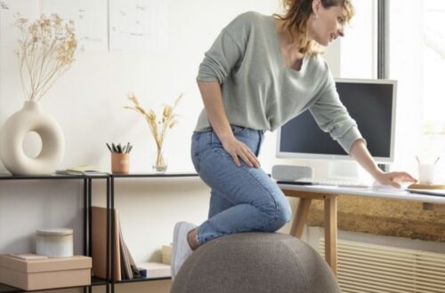 Woman resting knee on yoga ball chair whilst looking at computer on office desk.