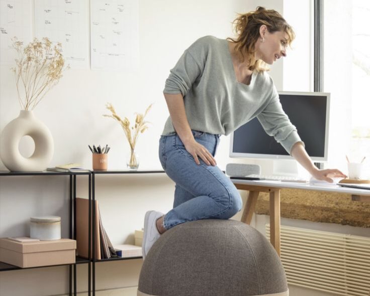 Woman resting knee on yoga ball chair whilst looking at computer on office desk.